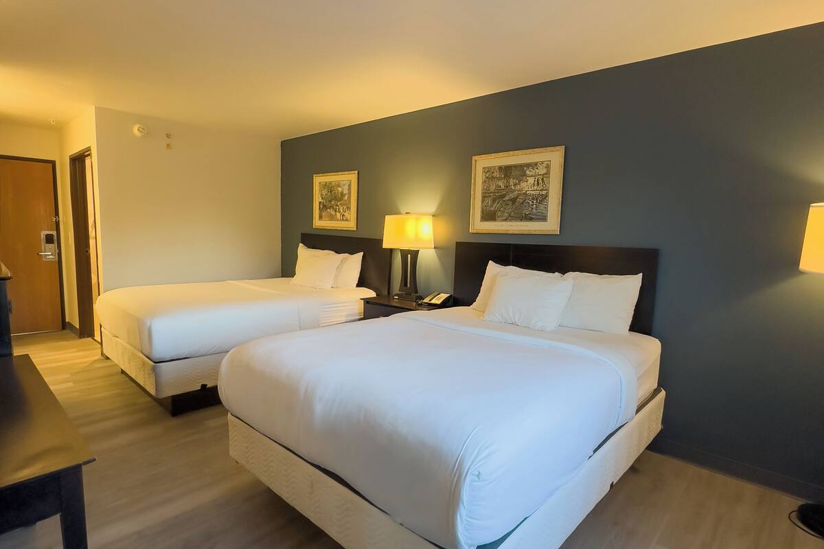 A hotel room with two beds, a desk, and a nightstand with a lamp.
