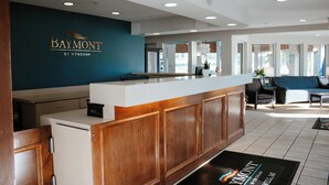 A reception area with a wooden reception desk, a blue wall with the Baymont logo, and a view of the lobby with seating and a plant. - Baymont by Wyndham Howell/Brighton (Howell)