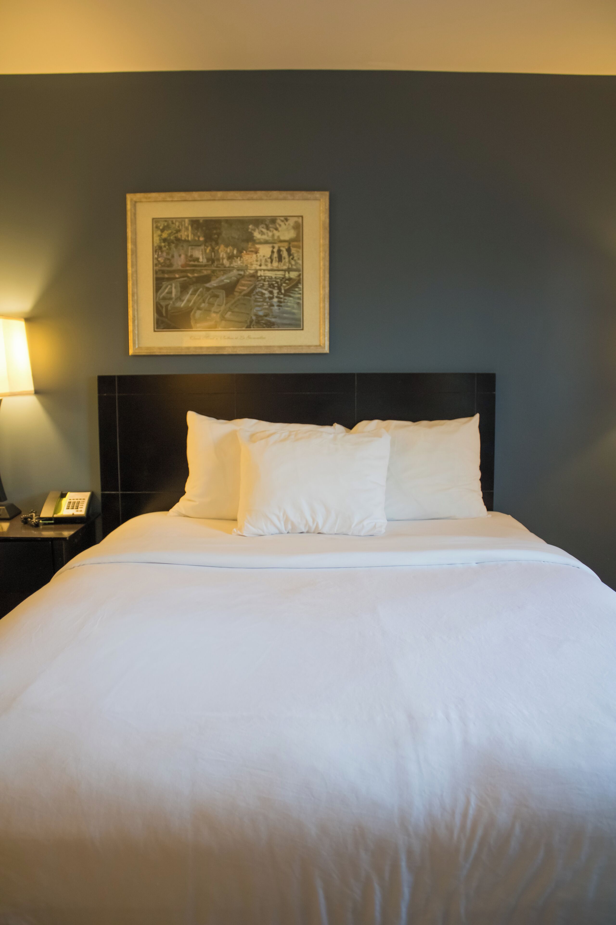 A neatly made bed with white linens, a dark headboard, and a framed picture on the wall above.
