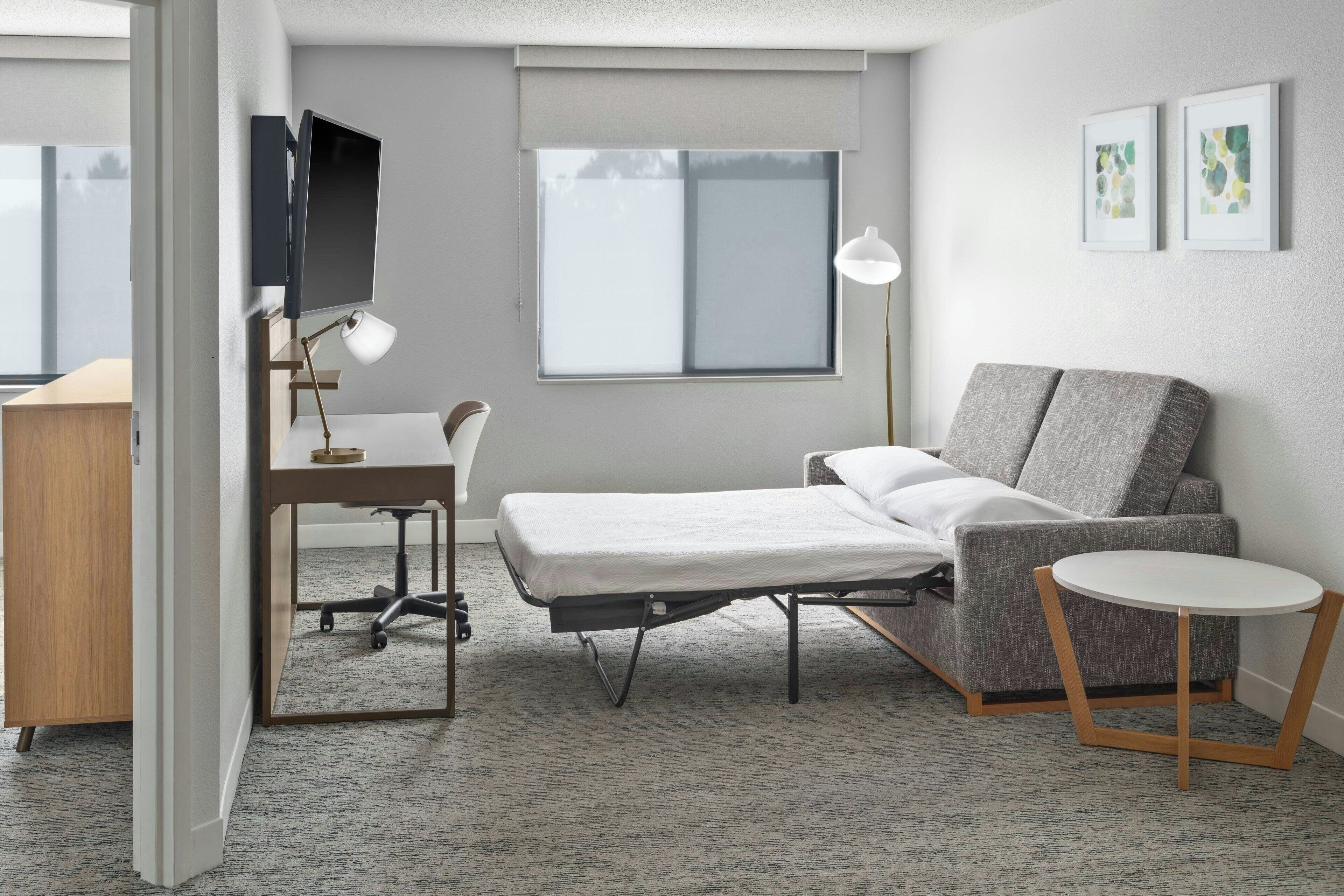 Quarto de hotel moderno com cama, escrivaninha com luminária, cadeira, mesa redonda pequena e televisão fixada na parede.