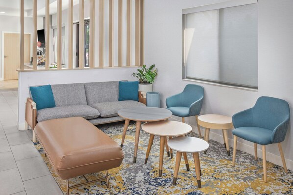 Un área de estar moderna con un sofá gris, sillones azules y un reposapiés marrón. Hay mesas de madera y una planta en maceta.