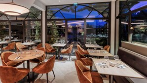Un restaurant avec des tables rondes et des fauteuils en velours, de grandes fenêtres donnant sur un paysage verdoyant et un décor intérieur moderne.