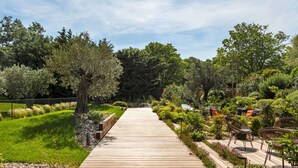 Un sentier en bois traverse un jardin où poussent différentes plantes et arbres, le tout étant entouré d’une clôture.