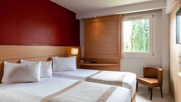 Quarto de hotel com duas camas, cabeceira de madeira, mesa de cabeceira, cadeira, janela com vista para árvores e uma parede de destaque vermelha.