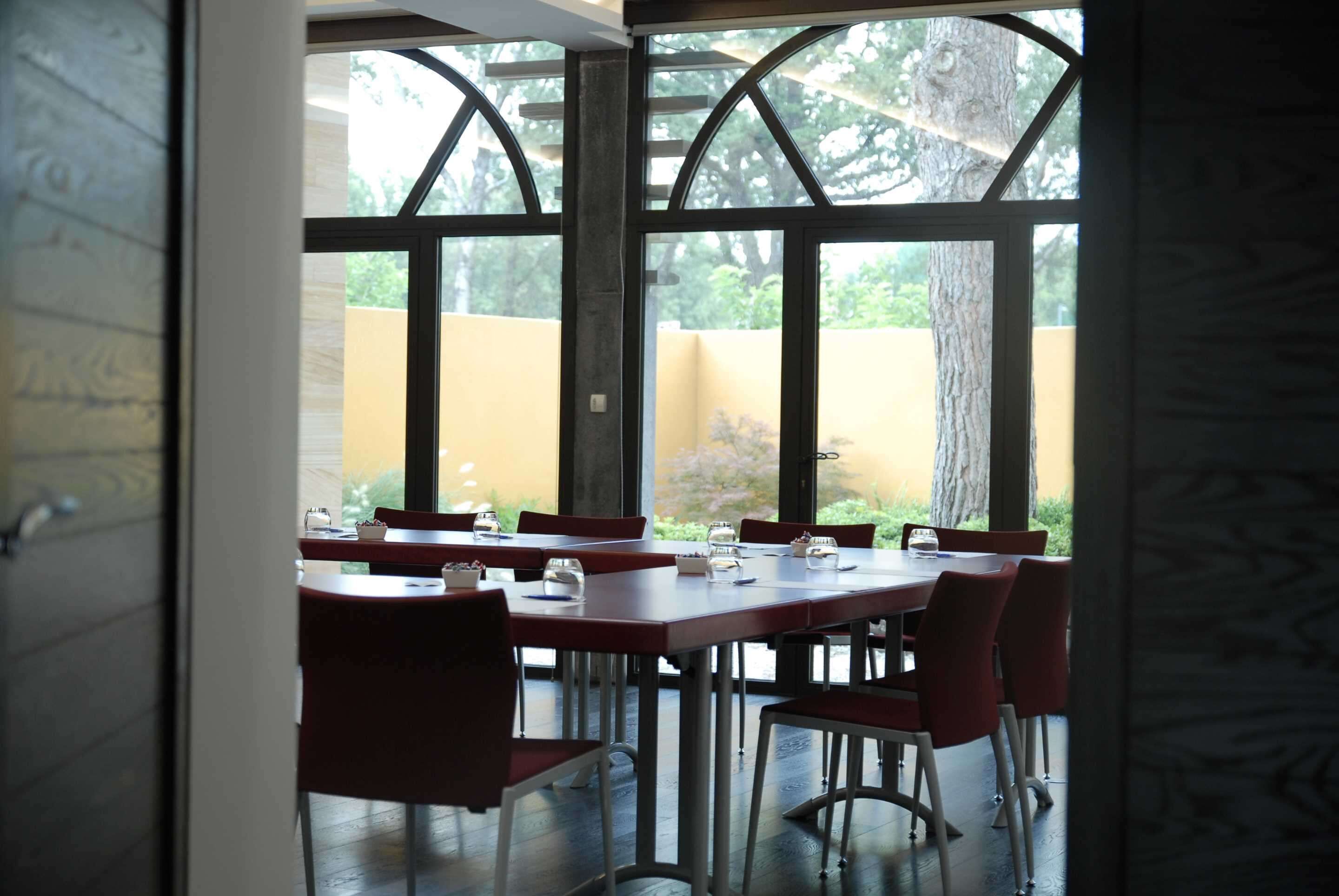 An indoor dining area with a large table and red chairs, large windows with a view of greenery outside.
