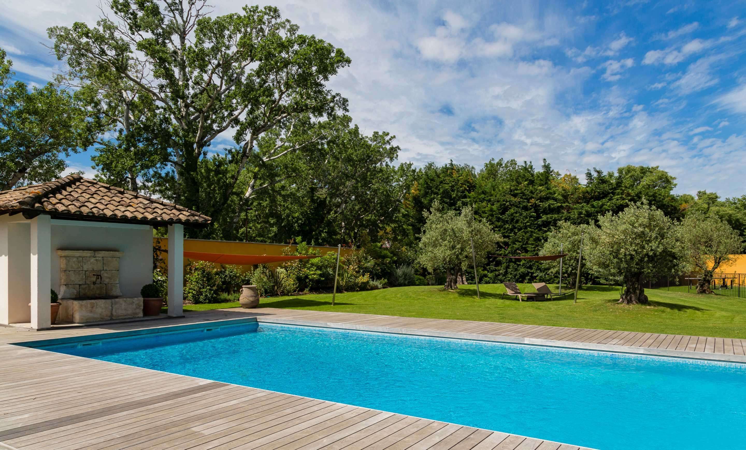 Ein Schwimmbad mit Holzterrasse, einem kleinen Pavillon und einer Rasenfläche mit Bäumen und einem Zaun.