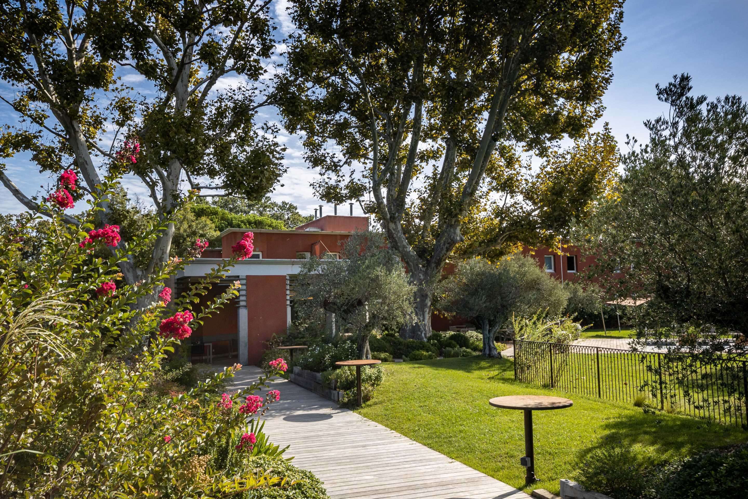 Un jardín con un sendero de madera, una mesa pequeña y un edificio con techo rojo.
