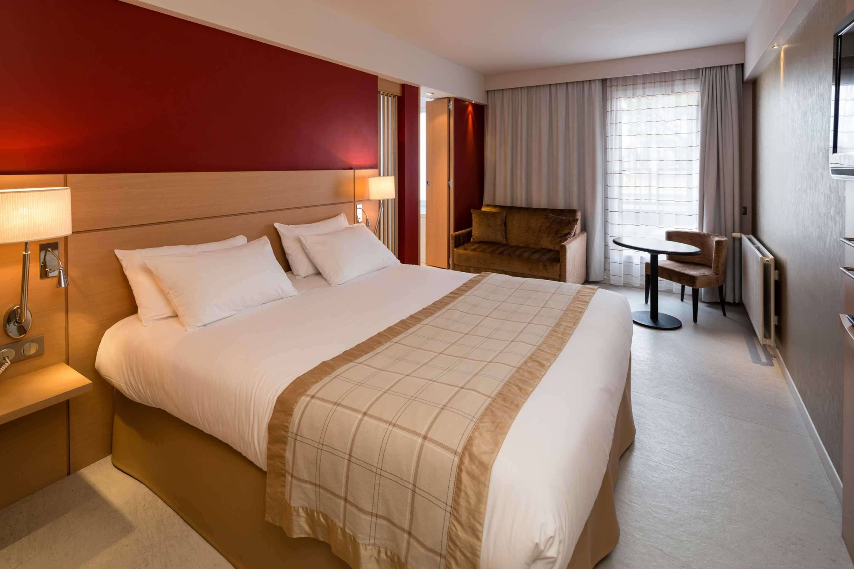 A hotel room with a large bed, a bedside table with a lamp, a sofa, a small table, and a window with curtains.