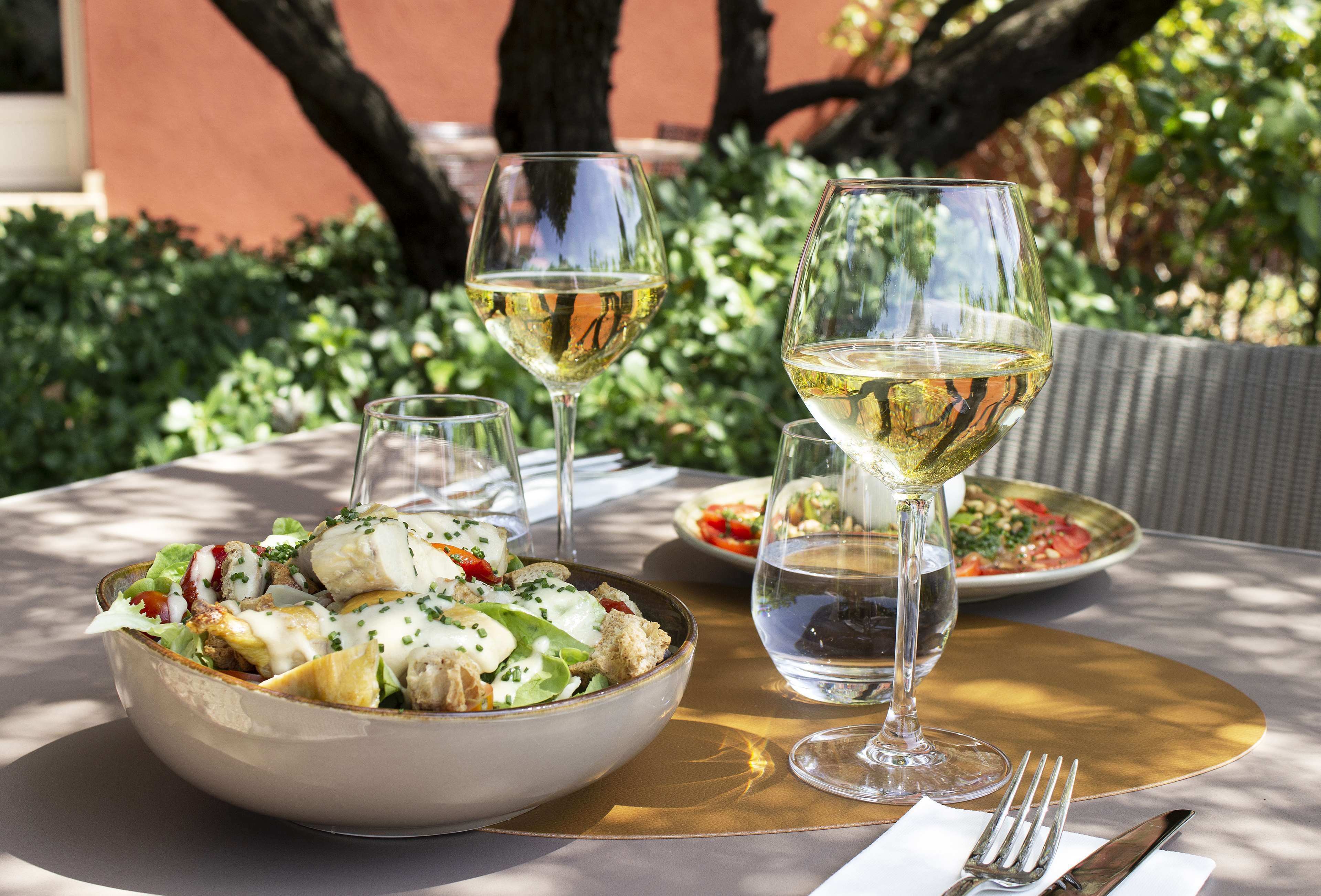 Une table dressée avec un saladier rempli de salade, deux verres de vin blanc et une fourchette et un couteau posés sur un set de table doré.