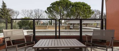 A balcony with a table and two chairs, overlooking a building and trees.
