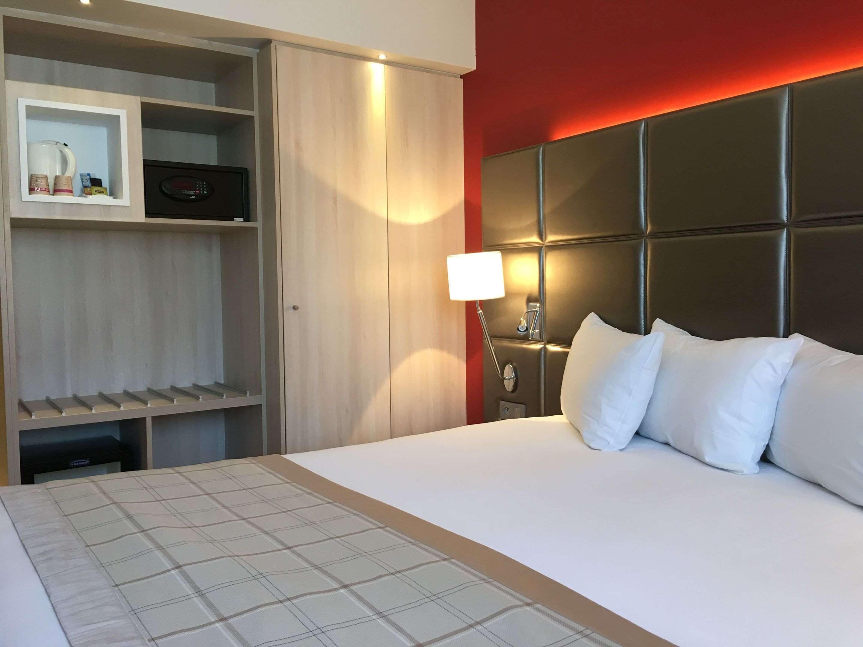 A hotel room with a bed, a wardrobe, a bedside table with a lamp, and a red accent wall.