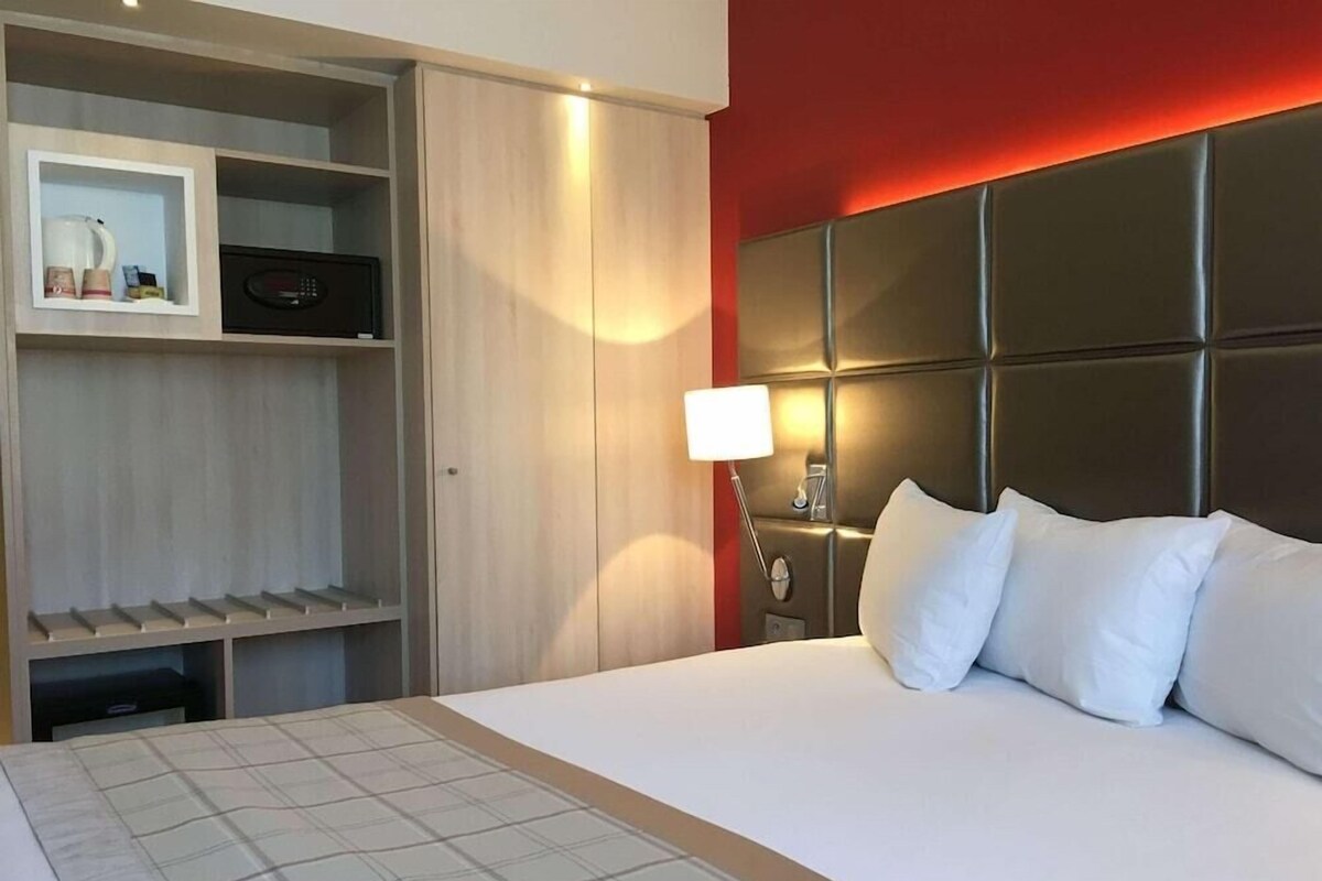 Una habitación de hotel con cama, mesita de noche, lámpara, armario y estante con objetos.