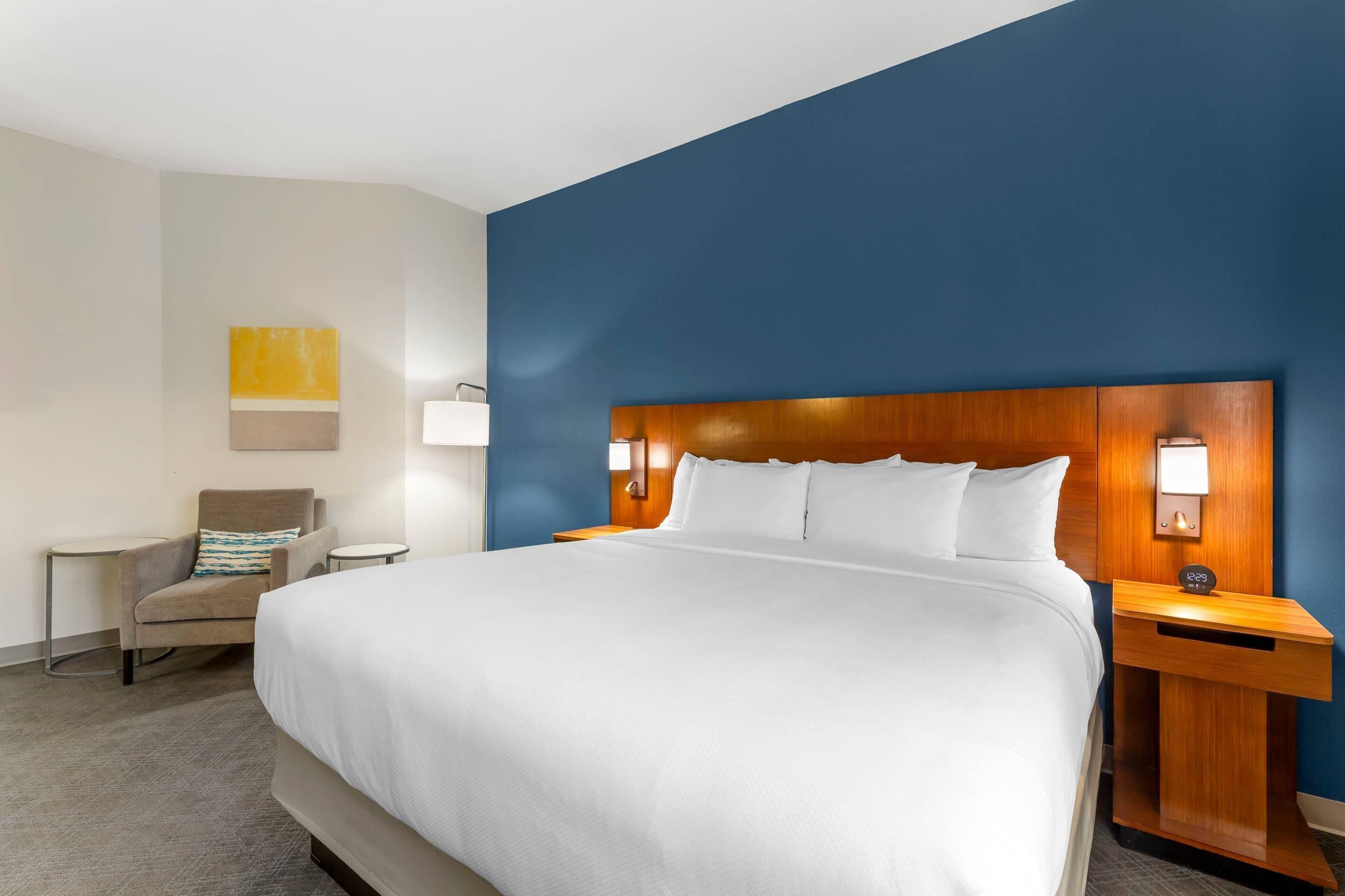 A hotel room with a large bed, a wooden headboard, bedside lamps, a chair, and a small table.