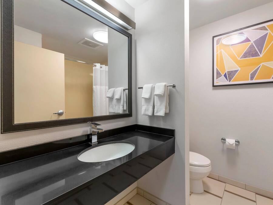 A modern bathroom with a black countertop, a white sink, a large mirror, and a toilet. There is a framed abstract art piece on the wall.