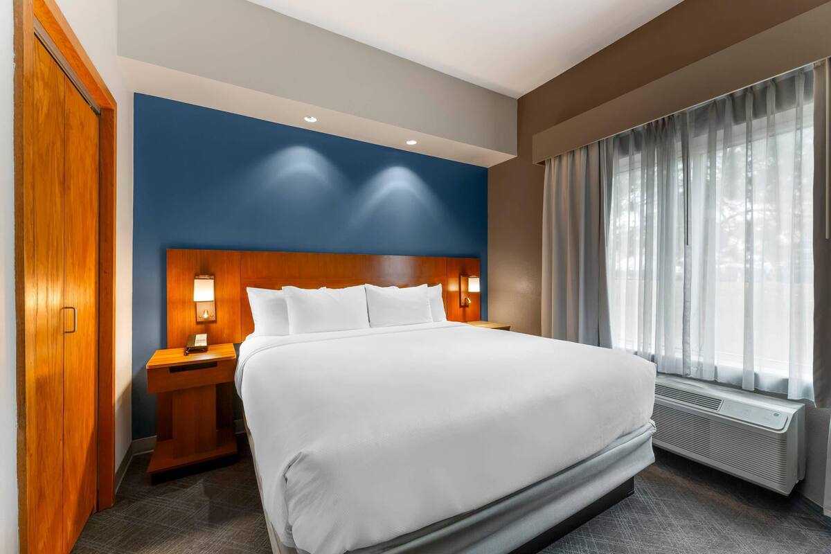 A hotel room with a large bed, a wooden headboard, a nightstand, a desk, a window with curtains, and a wall with recessed lighting.
