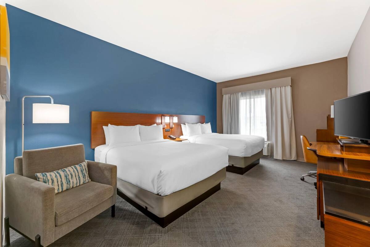 A hotel room with two beds, a desk, a chair, a TV, and a lamp.