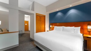 Habitación de hotel con una cama grande, cabecera de madera, escritorio con silla y una zona de recepción.