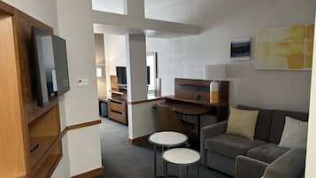 A modern hotel room with a sofa, two small tables, a TV, and a desk with a lamp.