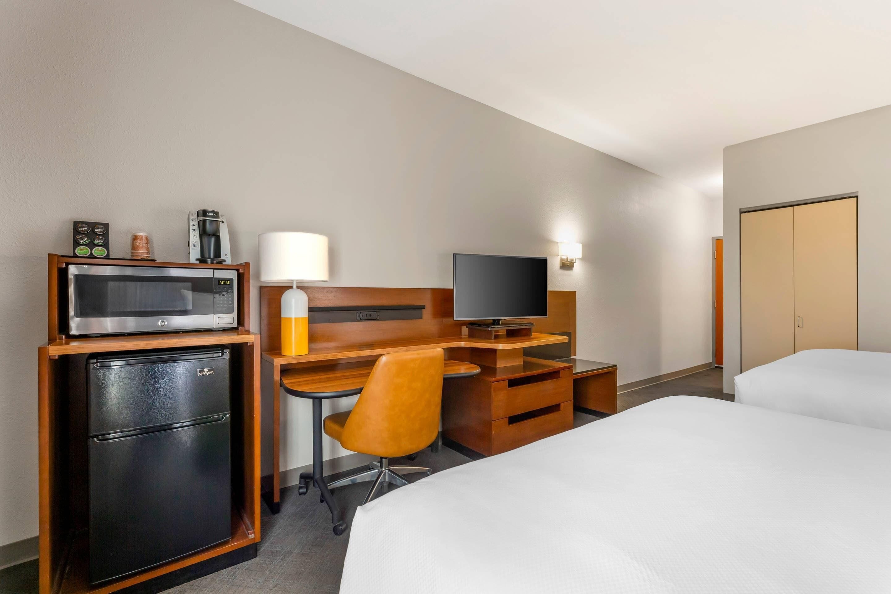 A hotel room with a bed, a desk with a computer, a microwave, a refrigerator, a TV, and a lamp.
