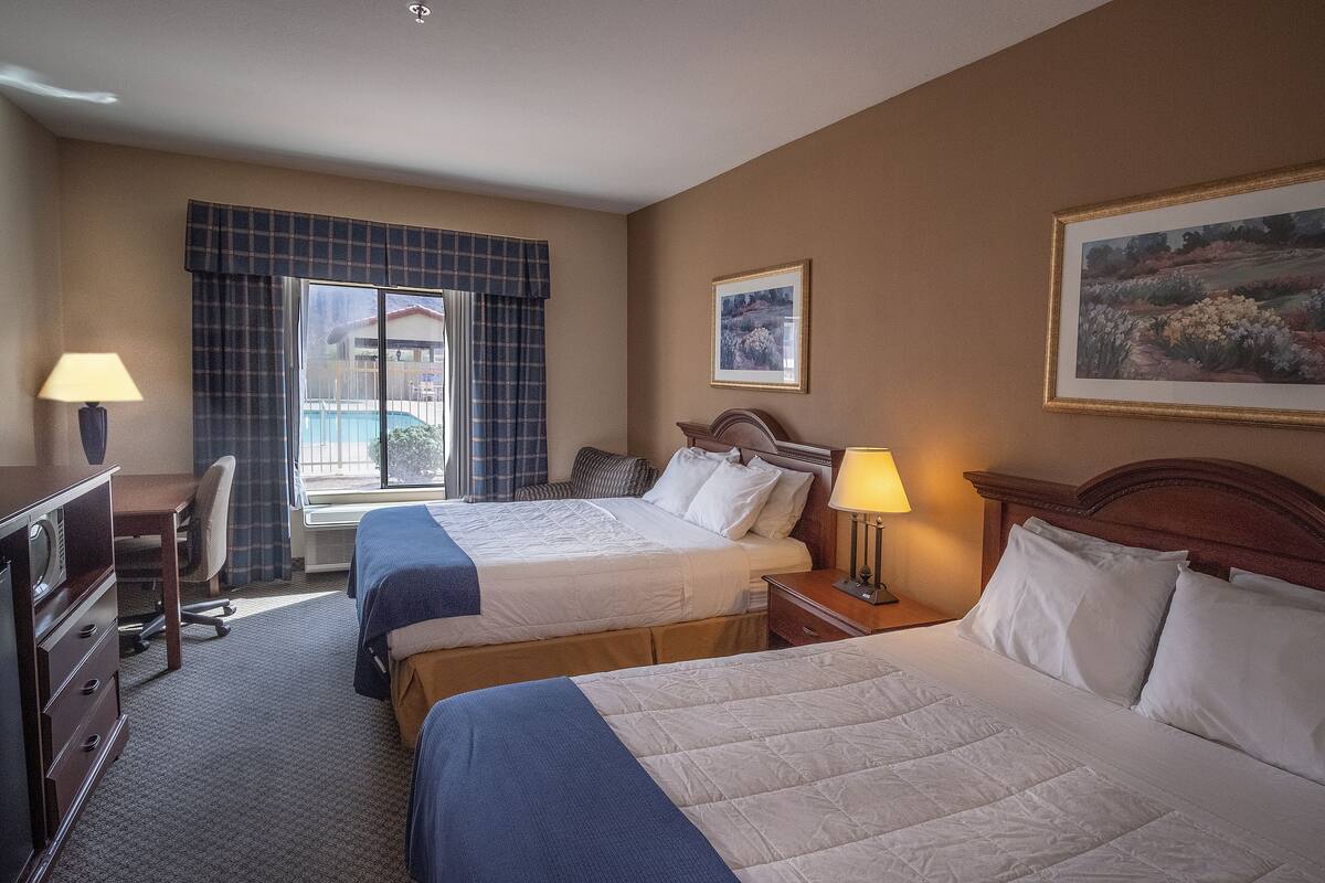 A hotel room with two beds, a desk, a chair, a lamp, and a window with a view of a building.