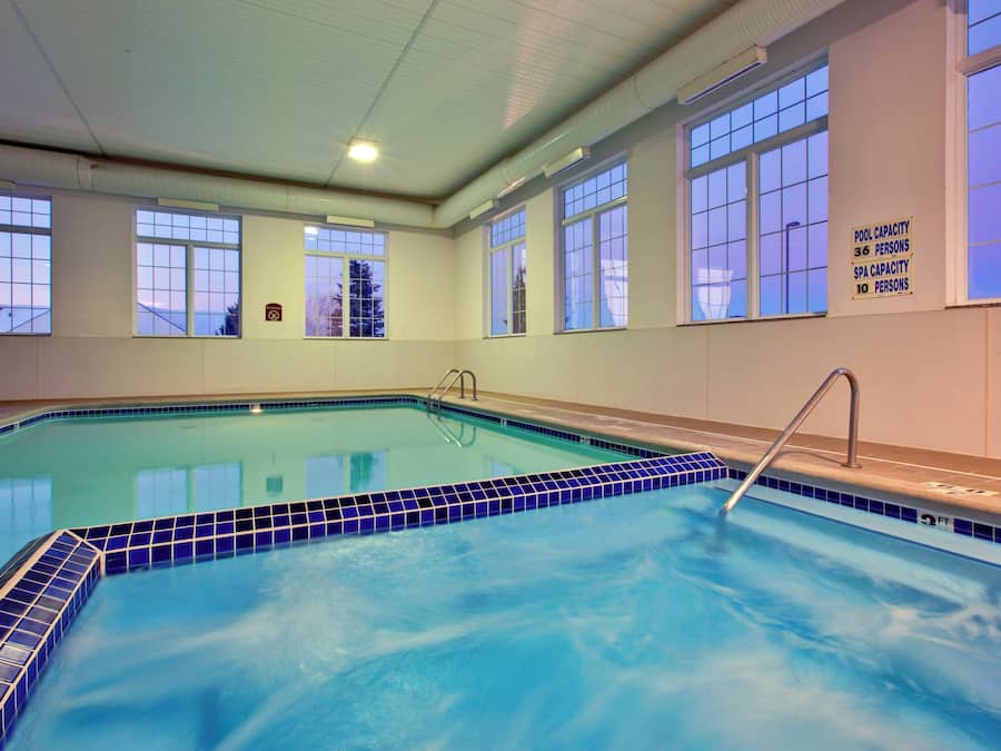 An indoor swimming pool with clear blue water, surrounded by windows, and a sign indicating pool and spa capacities.