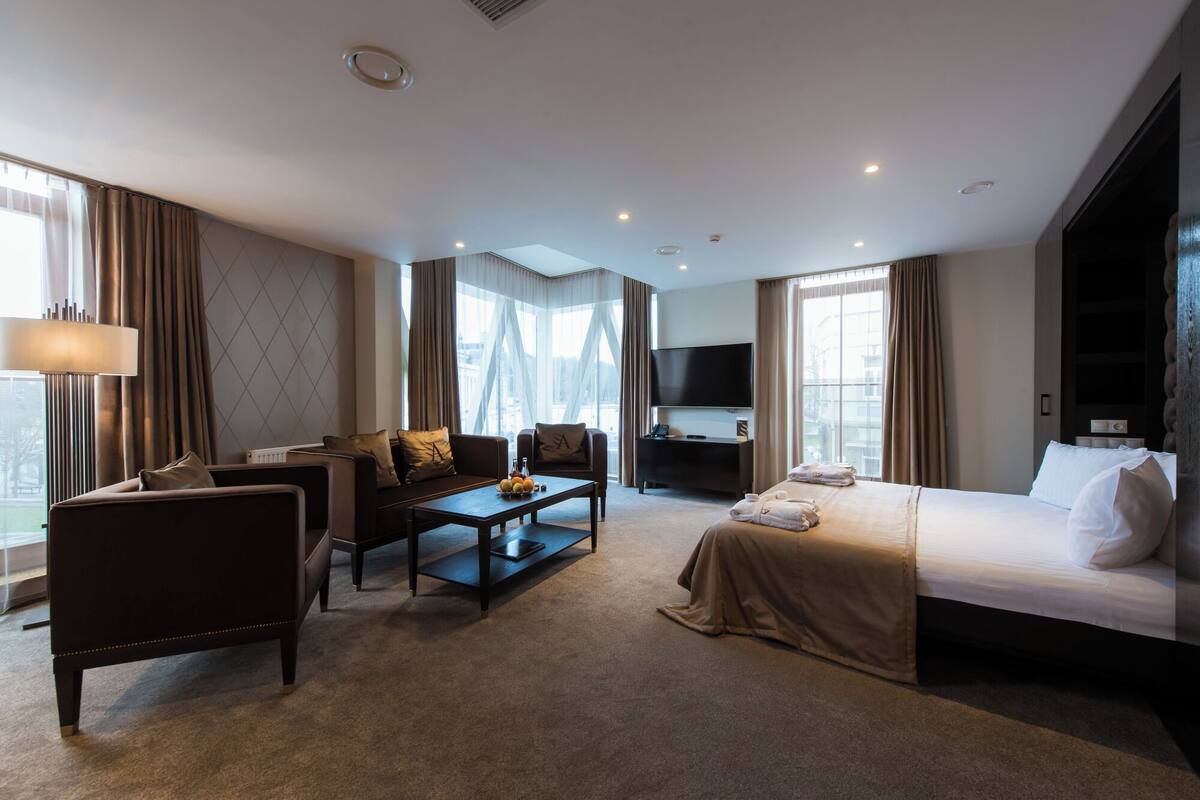Camera d'albergo con divano, tavolino, televisione, letto e ampie finestre con vista sulla città.