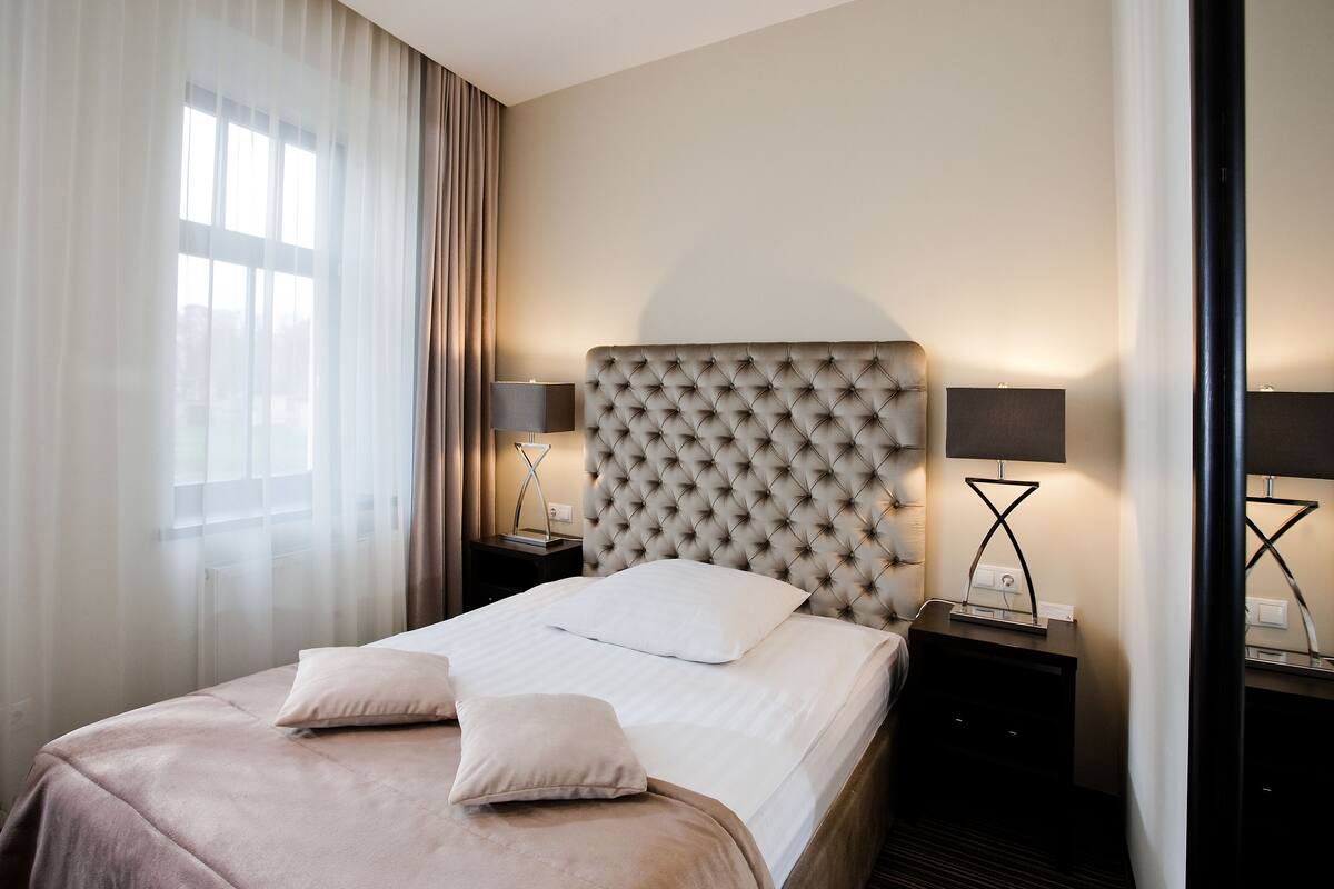 Una camera d'albergo con un letto grande, due comodini con lampade e una finestra con tende leggere.