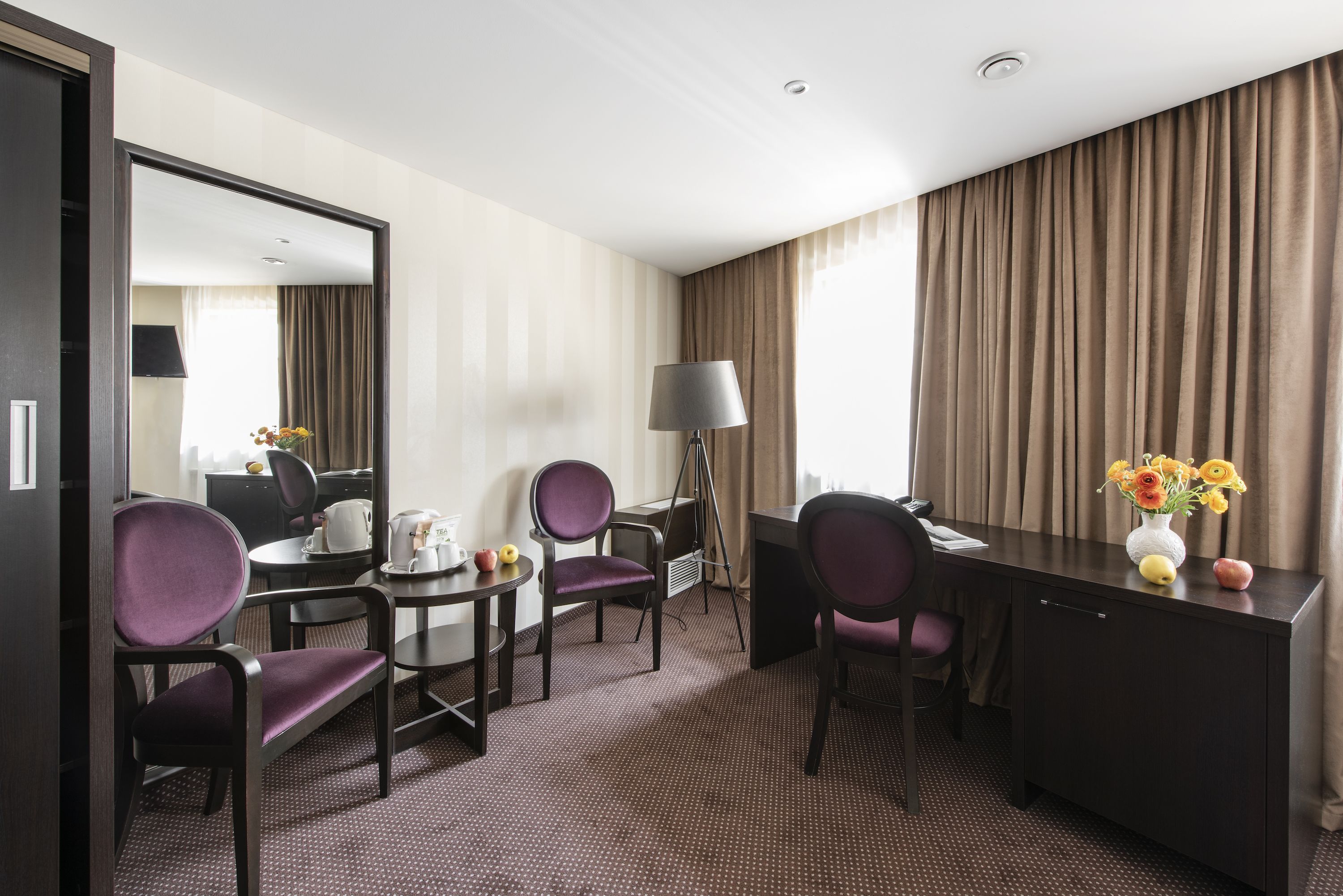 Habitación de hotel con escritorio, dos sillas, lámpara, espejo y un florero con flores.