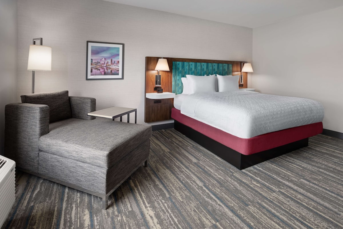 Habitación de hotel con cama, sofá, mesita de noche, lámpara y un cuadro en la pared.