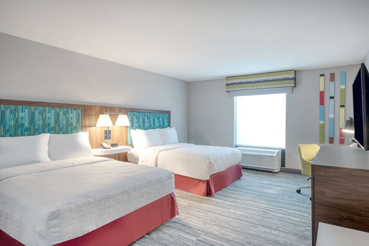 Una habitación de hotel con dos camas, un escritorio, una silla y un ventanal grande.