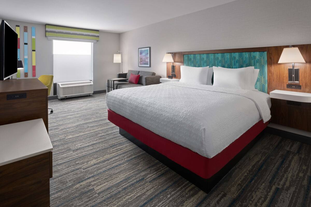 Habitación de hotel con una cama grande, un escritorio, una silla, un televisor, una ventana con persianas y una lámpara de pared.