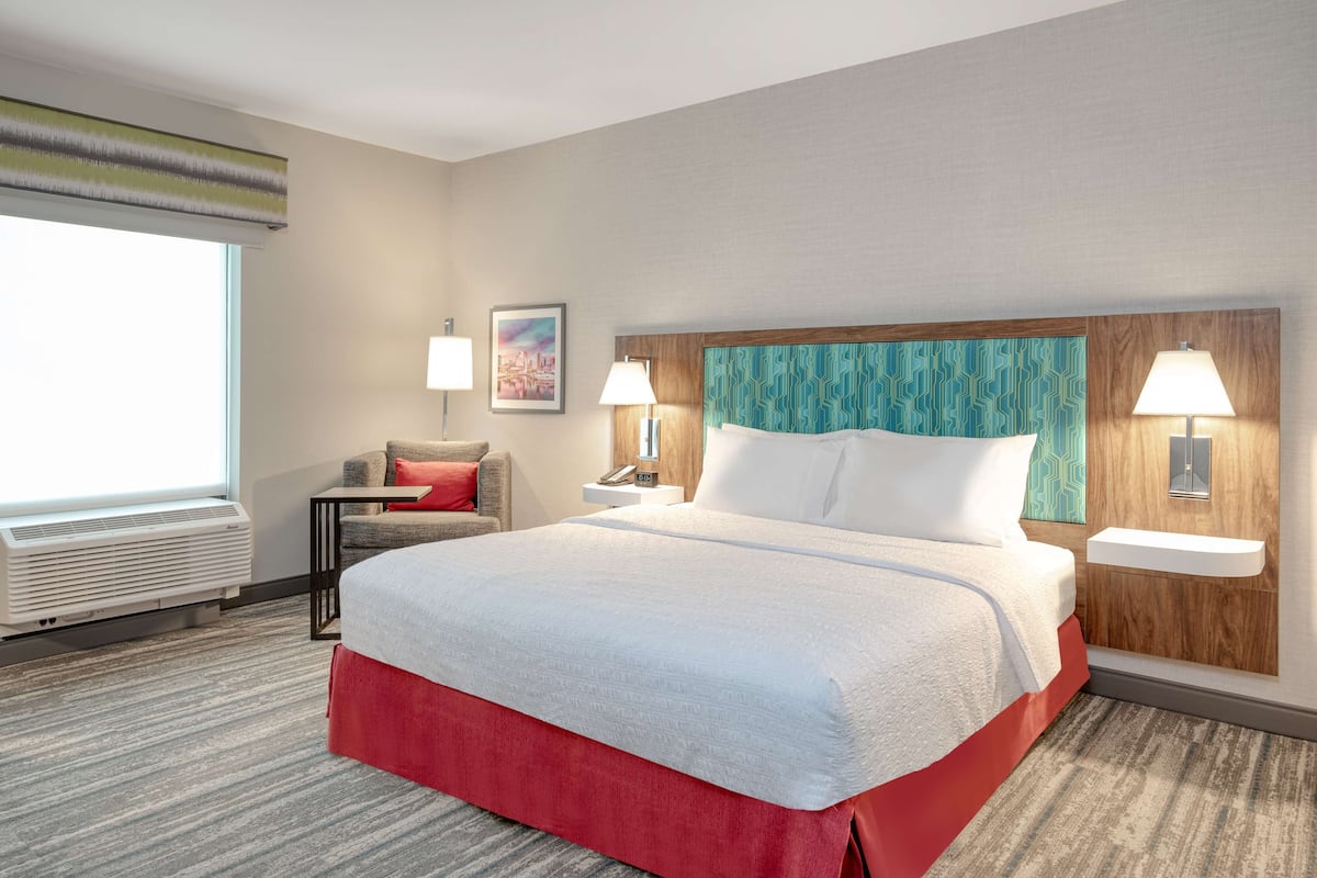 Habitación de hotel con una cama grande, dos mesitas de noche con lámparas, una silla, una ventana con persianas y un calefactor de pared.