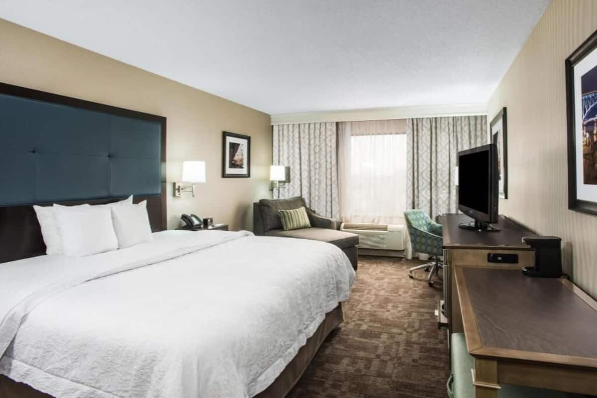 A hotel room with a large bed, a desk with a computer, a chair, a TV, and a window with curtains.