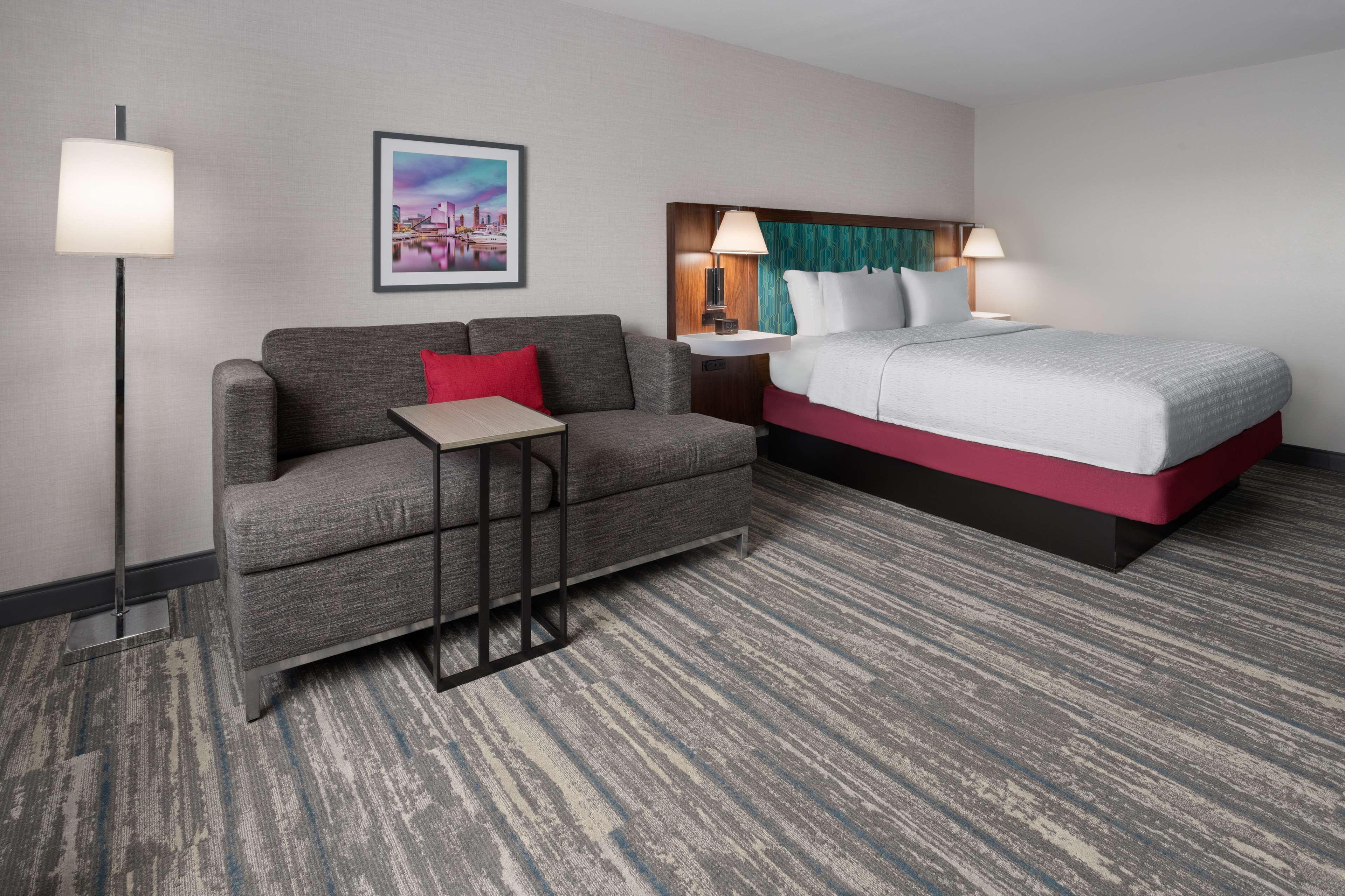 A hotel room with a grey sofa, a small table, a bed with red and white bedding, a nightstand with a lamp, and a framed cityscape picture on the wall.