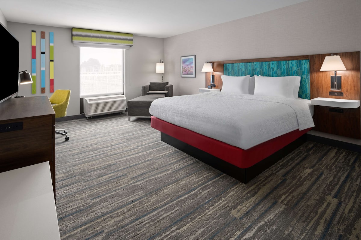 Habitación de hotel con una cama grande, un escritorio, una silla, un ventanal con vistas y una pared decorada con colores.