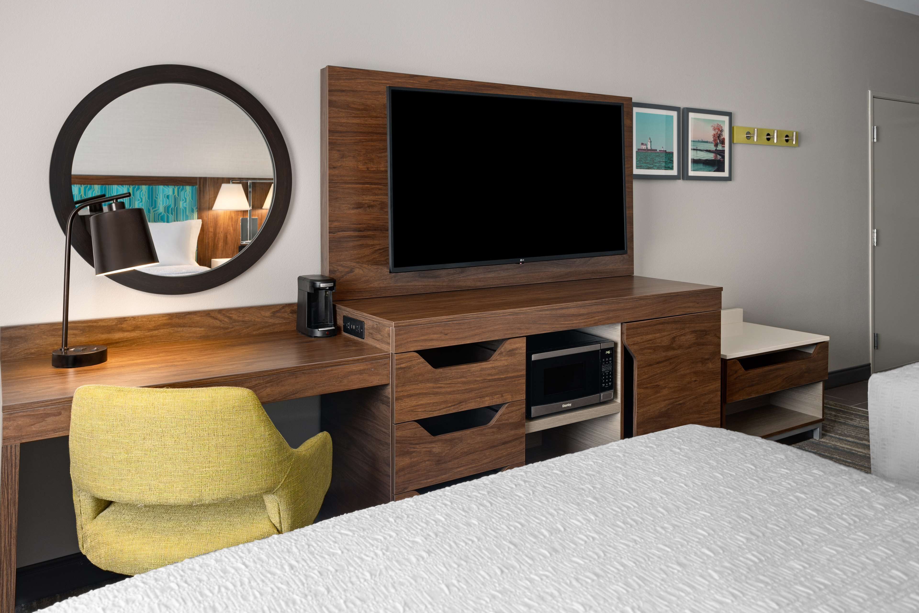 A hotel room with a large flat-screen TV, a wooden desk, a yellow chair, a bed with a white textured cover, and a round mirror on the wall.