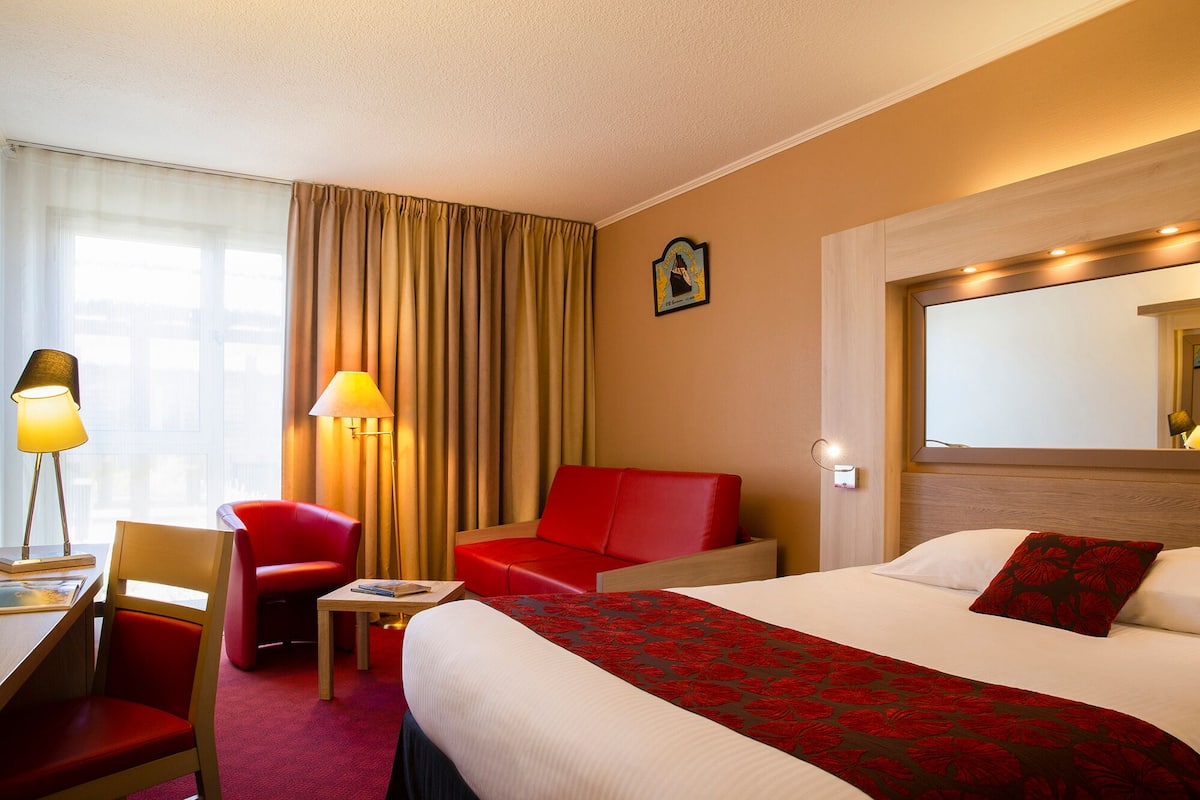 A hotel room with a bed, a desk with a chair, a red armchair, a small table, and a lamp.