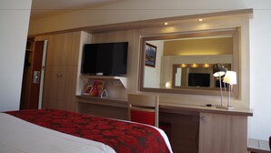 A hotel room with a bed, a TV, a desk with a lamp, a mirror, and a shelf with books and a magazine.