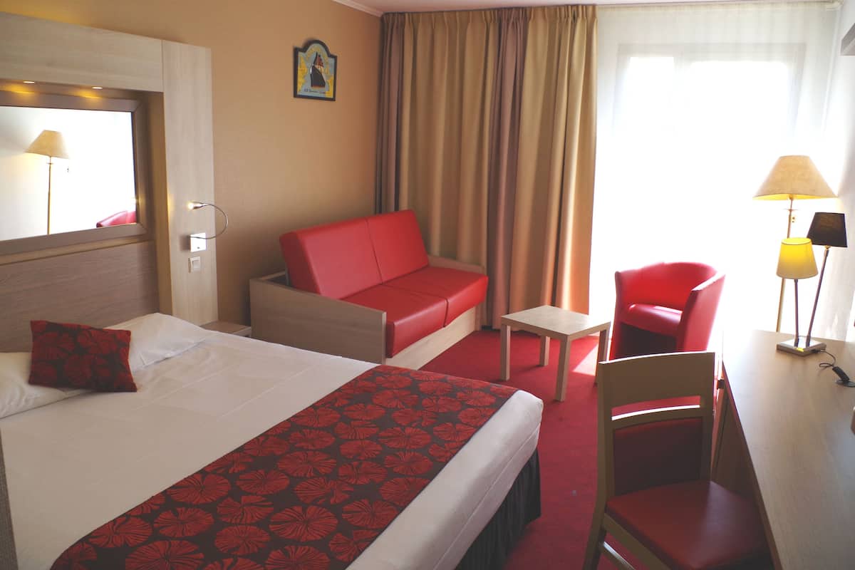 A hotel room with a bed, a red sofa, a red chair, a small table, and a desk with a lamp.