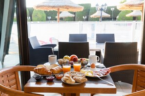 Una mesa de desayuno con croissants, fruta y jugo, con vistas a un jardín cuidado y sombrillas.