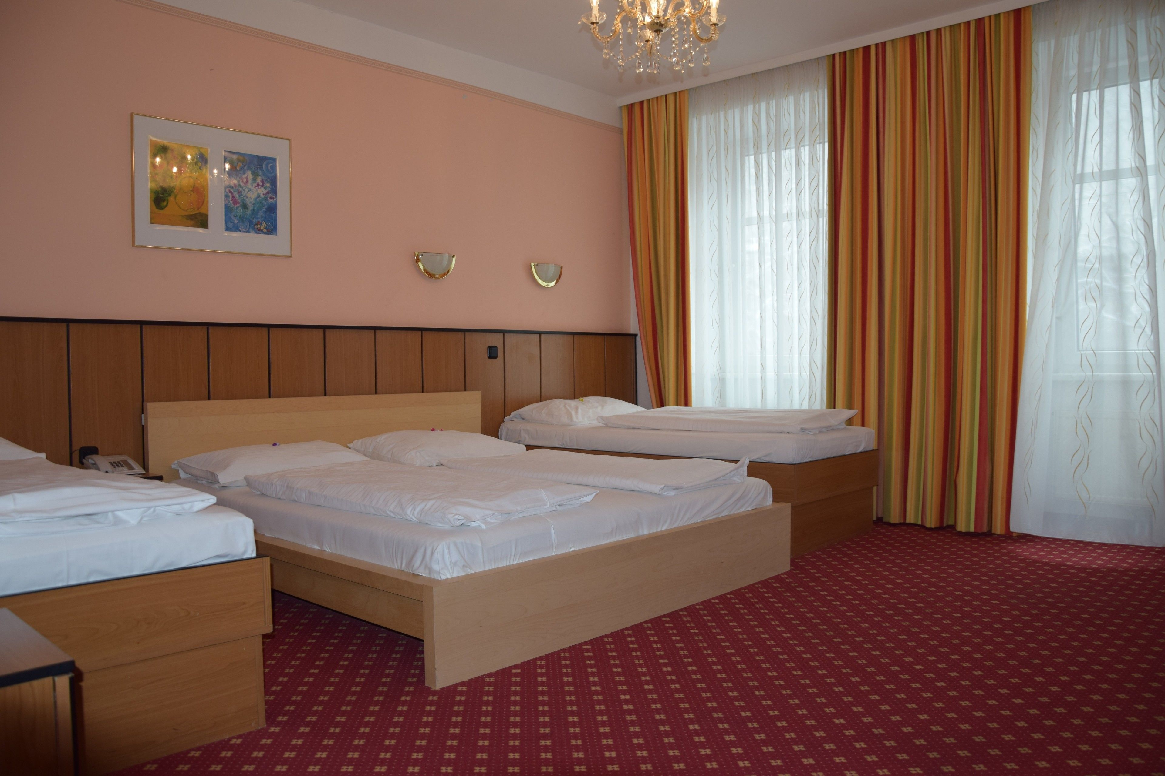 Una camera d'albergo con tre letti, una testiera in legno, una moquette rossa a motivi e un'ampia finestra con tende a righe.
