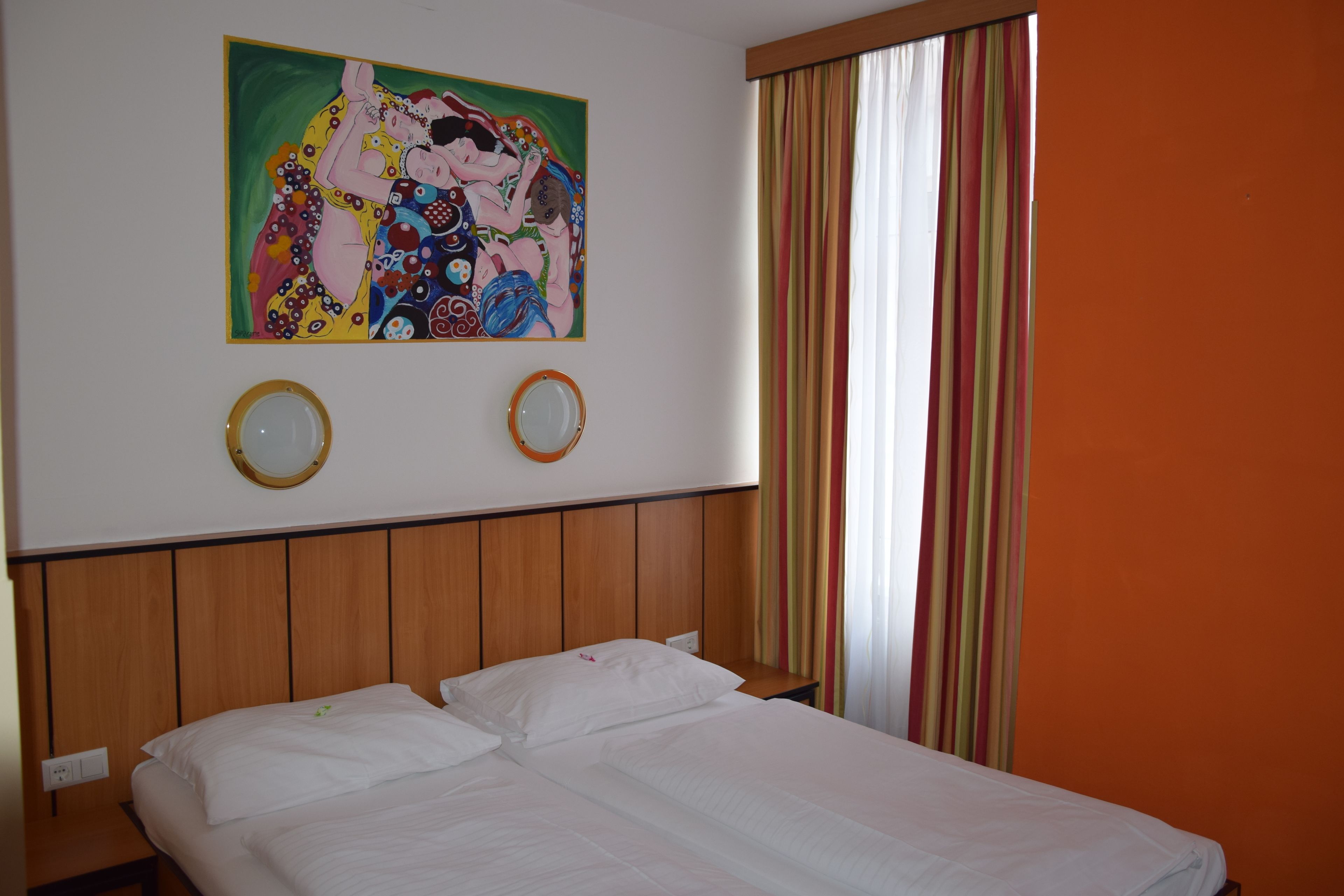 Une chambre d’hôtel avec une tête de lit en bois, deux miroirs ronds, un tableau au mur et un lit recouvert de draps blancs.