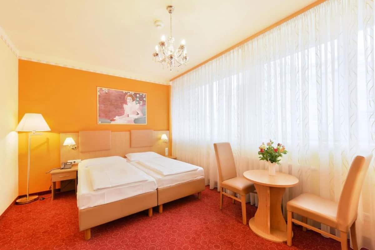 A hotel room with two beds, a desk, a chair, a small table with flowers, and a chandelier.