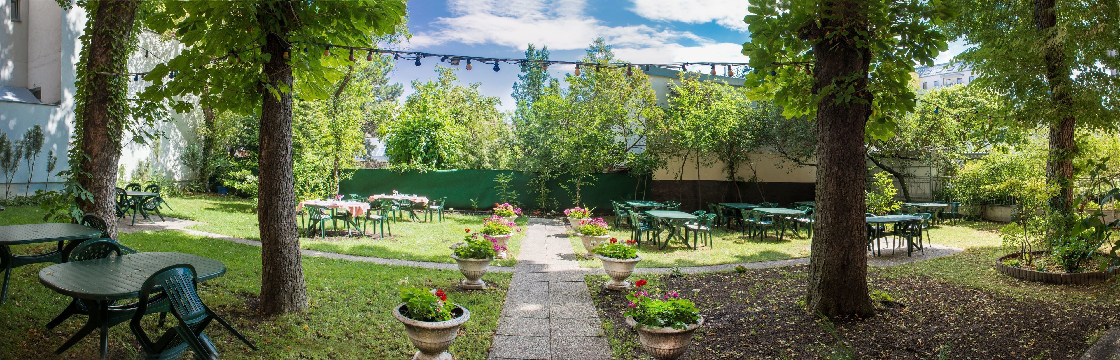 An outdoor patio with green tables and chairs, surrounded by trees and potted plants.