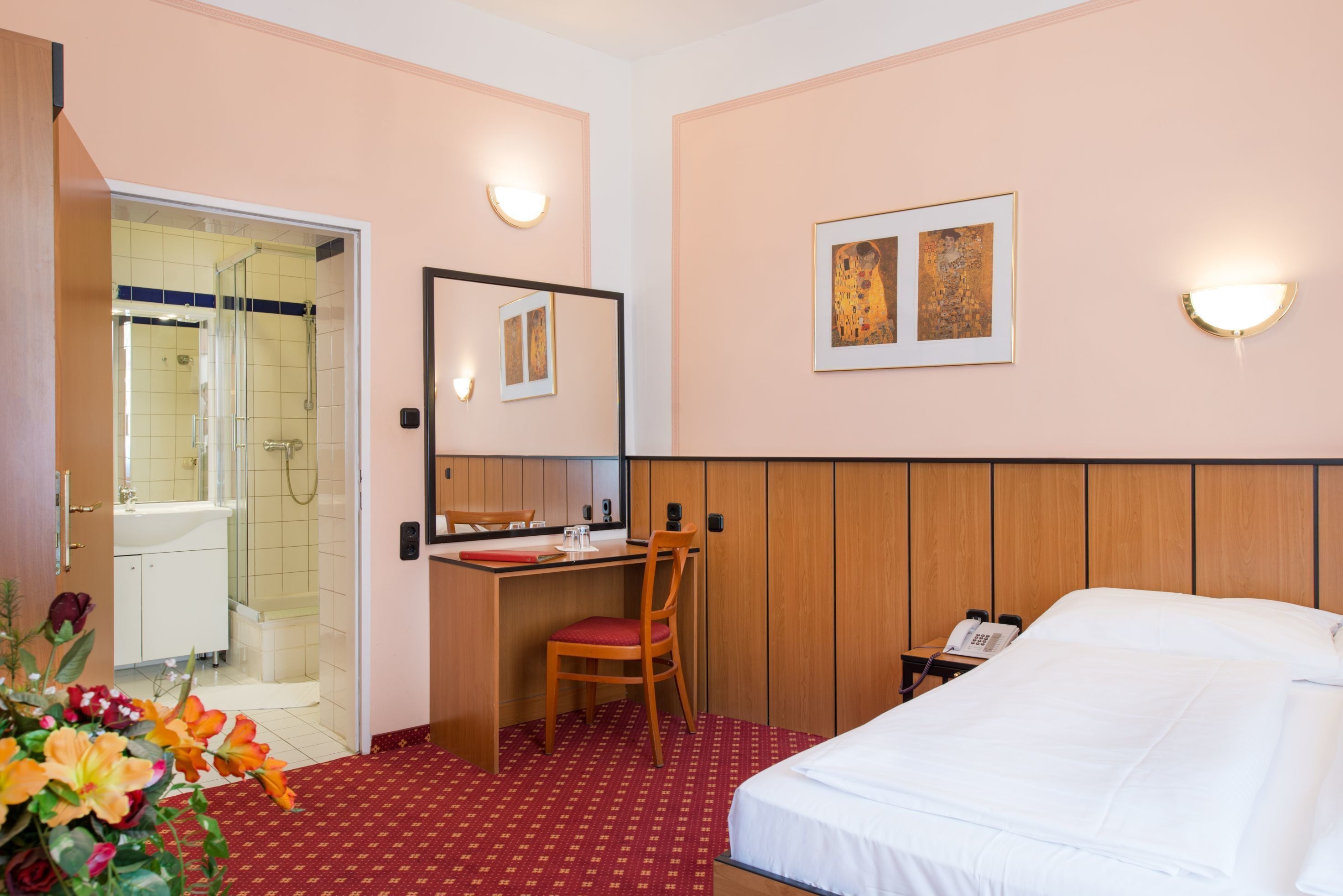 A hotel room with a bed, a desk with a chair, a mirror, a phone, and a shower area.