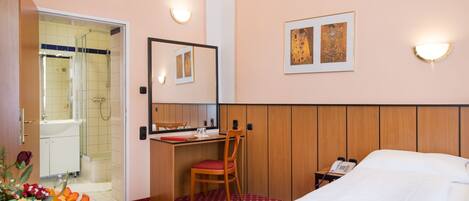 A hotel room with a bed, a desk with a chair, a mirror, a phone, and a shower area.