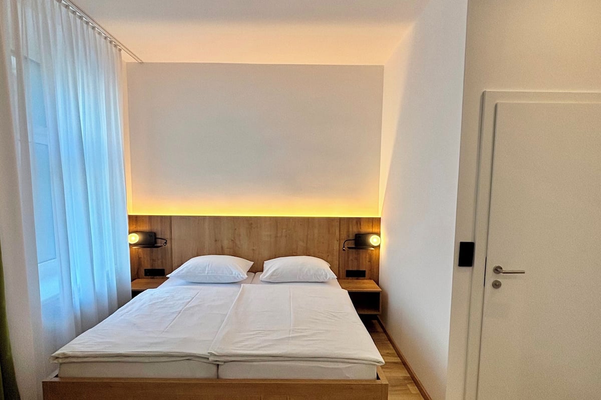 Ein Doppelbett mit zwei Nachttischen, einem Holz-Kopfteil und einer Tür rechts.