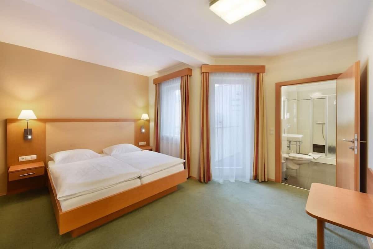 Ein Hotelzimmer mit Doppelbett, Nachttischlampen, einem kleinen Tisch, einem Spiegel und einem durch eine Glastür einsehbaren Badezimmer.
