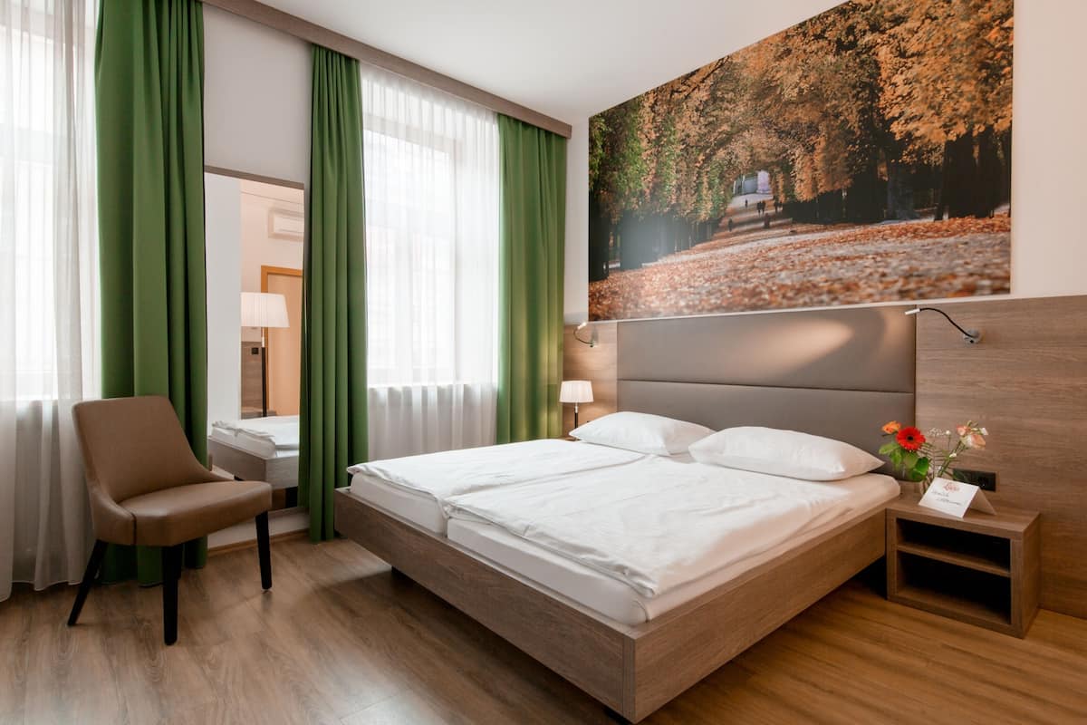 Ein Hotelzimmer mit einem großen Bett, einem Sessel, einem Nachttisch mit Lampe und einem Gemälde einer Waldlandschaft.