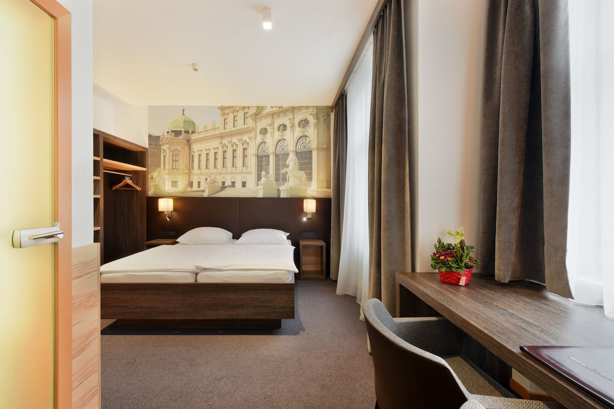 Ein Hotelzimmer mit Bett, Nachttisch, Lampe und Blick auf ein Gebäude durch das Fenster.